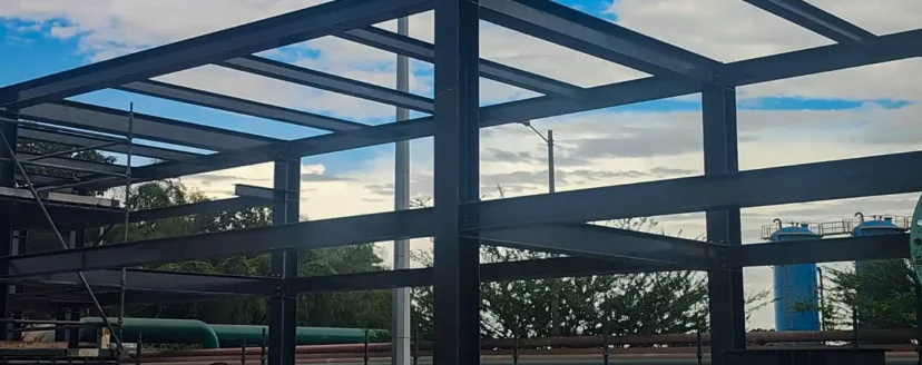 Infraestructura metálica a cielo abierto con armazón de acero oscuro, parte de un proyecto de obras civiles de GAPAC, destacando materiales y estructuras elaboradas por la empresa. Al fondo se aprecian tuberías industriales y un tanque, bajo un cielo parcialmente nublado.