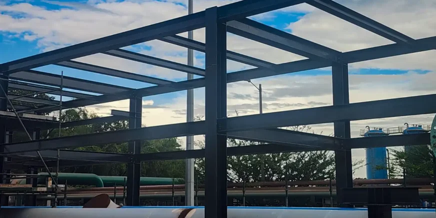 Infraestructura metálica a cielo abierto con armazón de acero oscuro, parte de un proyecto de obras civiles de GAPAC, destacando materiales y estructuras elaboradas por la empresa. Al fondo se aprecian tuberías industriales y un tanque, bajo un cielo parcialmente nublado.